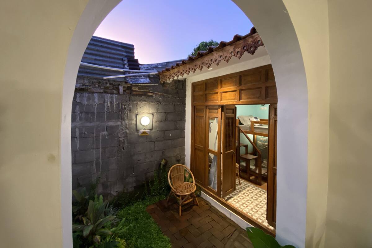 A cozy courtyard with a wooden bench, a wall-mounted light, and a view through an archway into a room with a patterned floor and wooden furniture.