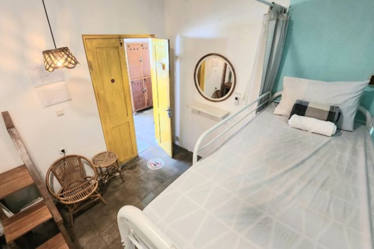 Ein Zimmer mit einem Bett, einem Nachttisch, einem Spiegel, einer Holztür und zwei Korbstühlen.