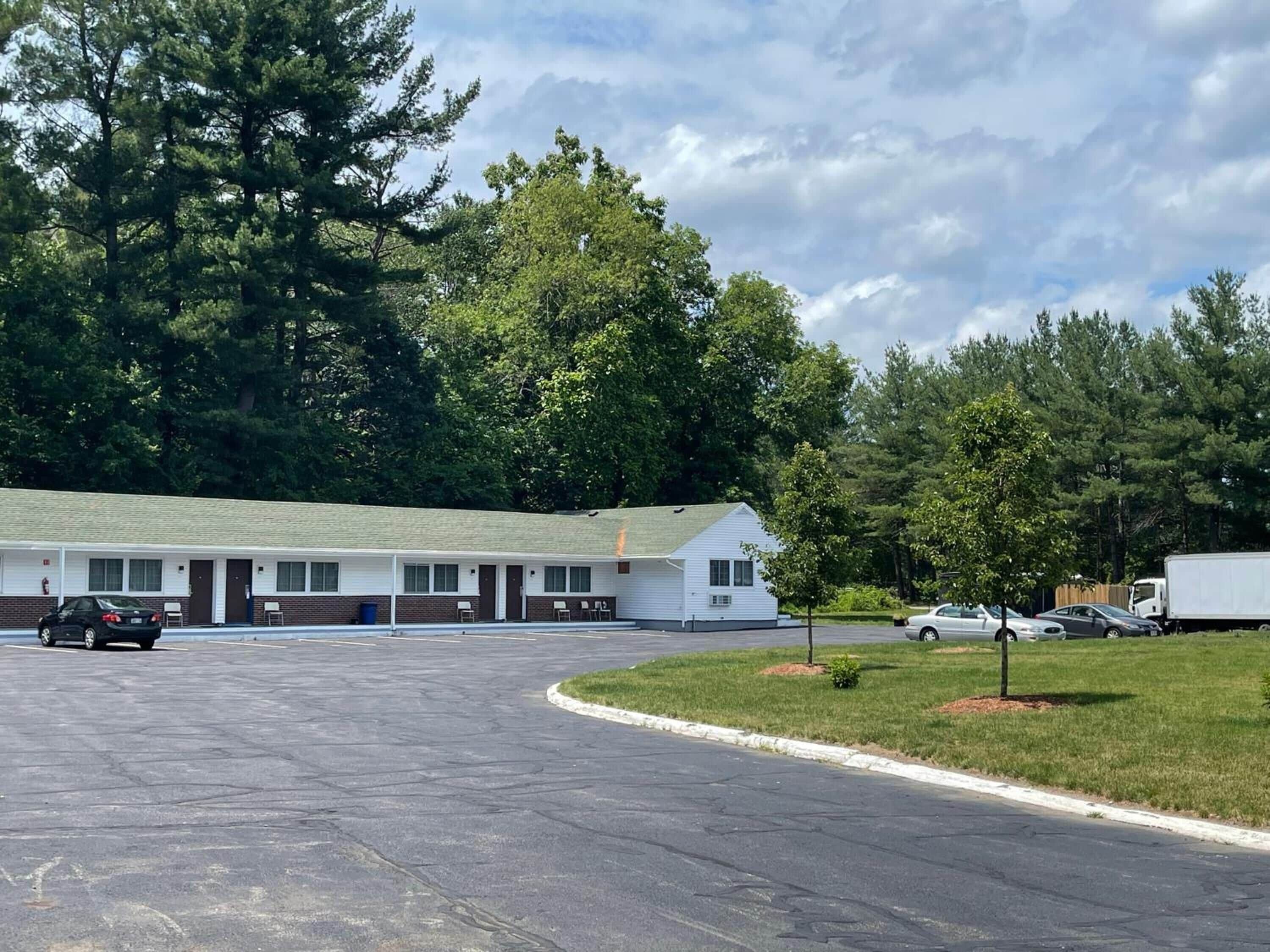 A motel with a row of rooms, a parking lot, and a grassy area with trees.