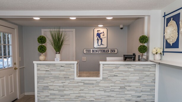 A reception area with a stone counter, a sign reading "The Minuteman Inn," and two potted plants. - Howard Johnson by Wyndham Acton (Acton)