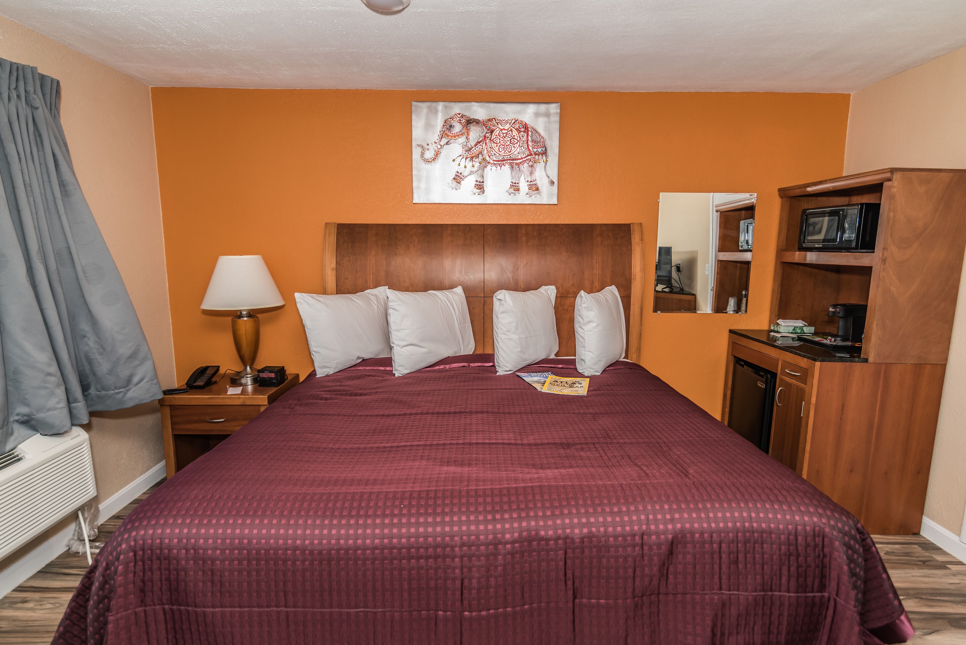 Habitación de hotel con una cama grande, cabecera de madera, mesita de noche con lámpara, espejo, armario y una obra de arte con elefantes en la pared.