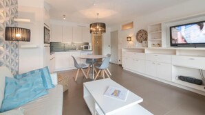 Uma sala de estar moderna com um sofá, uma mesa de centro e um espaço de jantar com cadeiras. Há uma cozinha com armários brancos e um micro-ondas. Uma televisão está fixada na parede, e um relógio é visível.