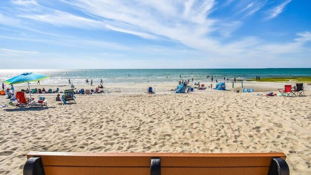 A beach with a wooden bench in the foreground, people walking, and beach chairs and umbrellas.