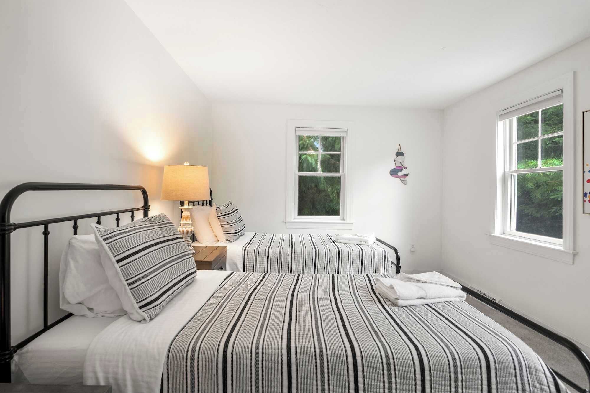 Un dormitorio con dos camas, una mesita de noche, una lámpara y dos ventanas.