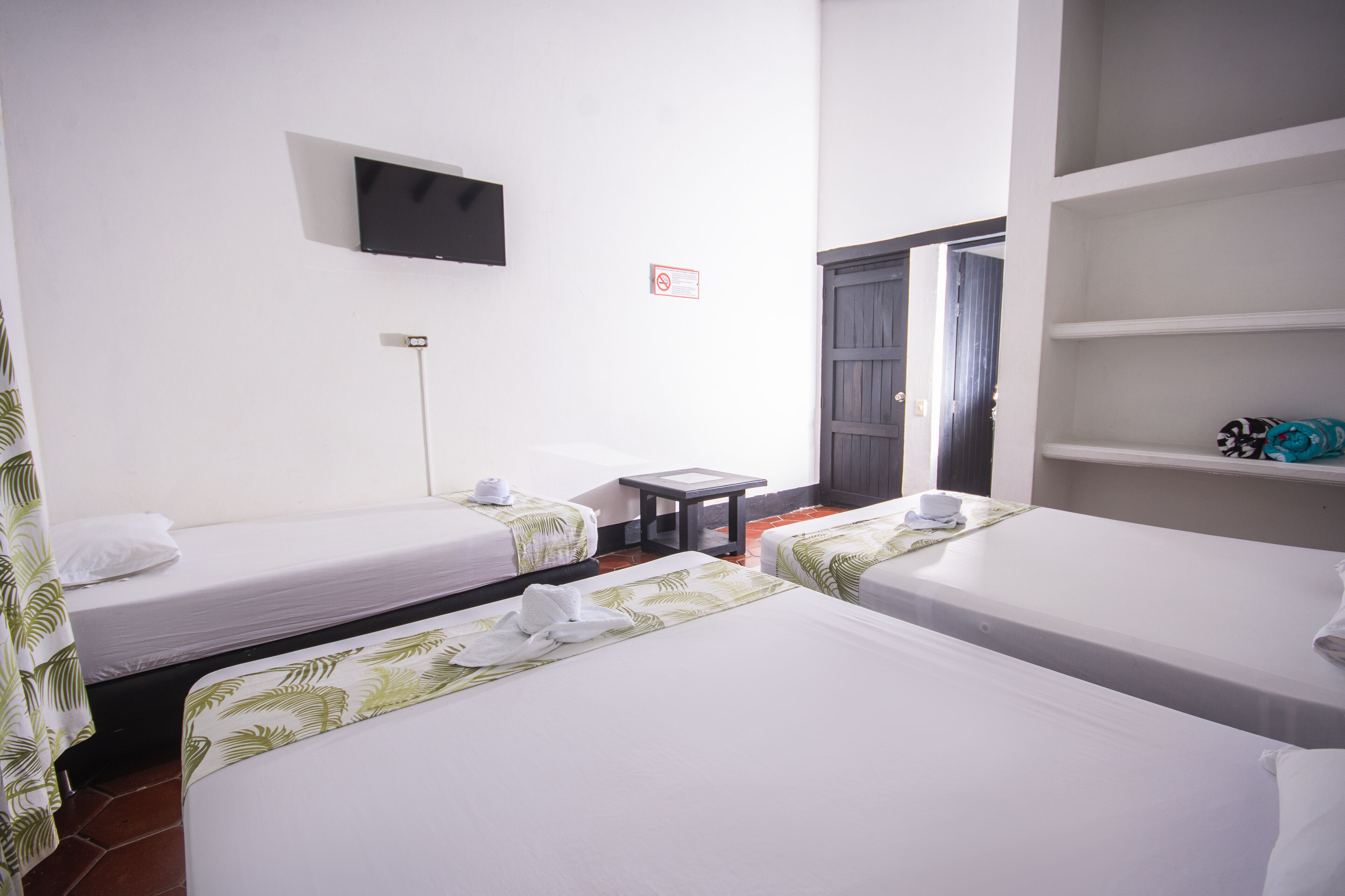 A hotel room with two single beds, a TV mounted on the wall, a small table, and a door leading to another room.