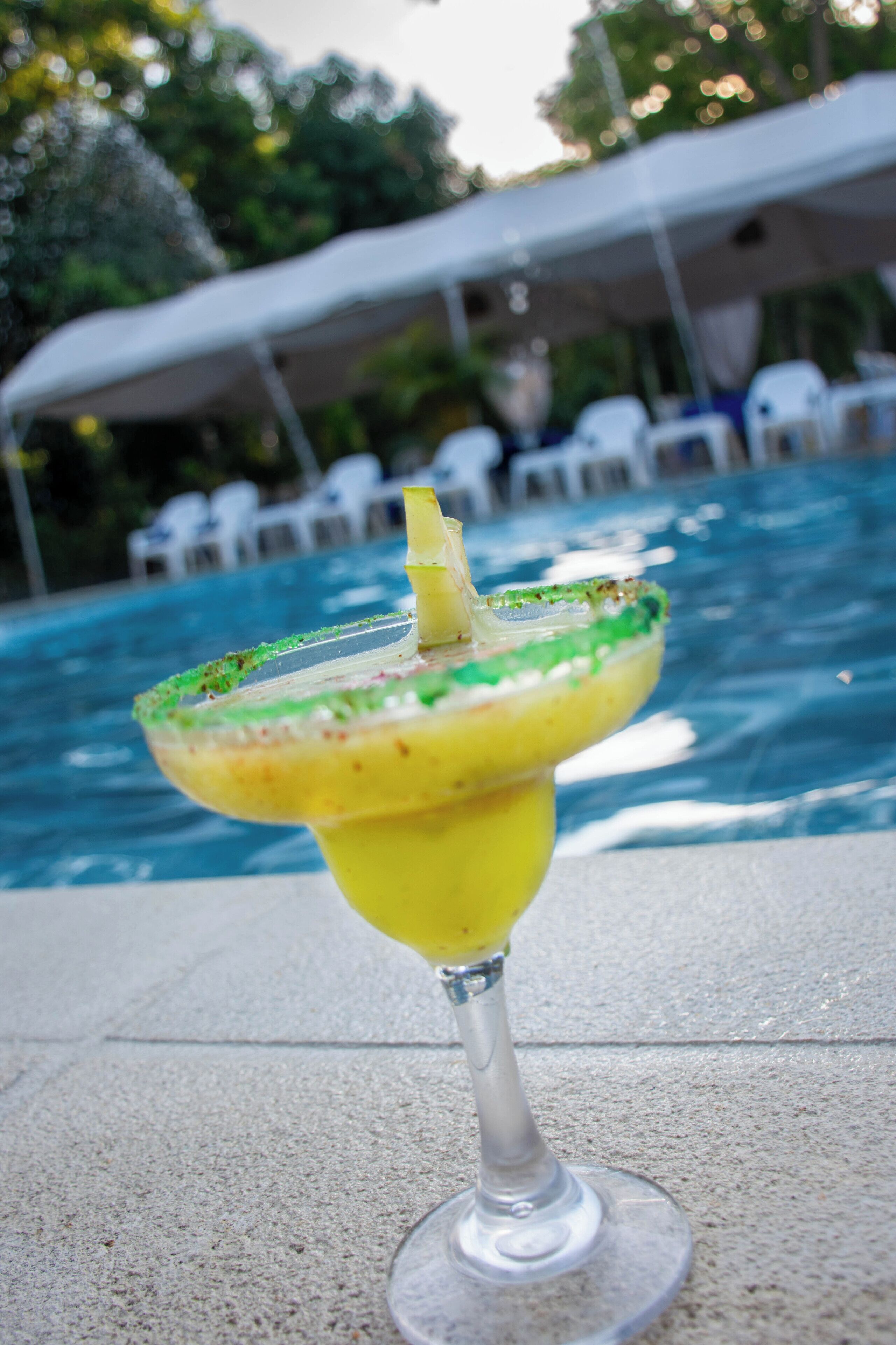Un cóctel margarita en un vaso con borde de sal, sobre una piscina con una pérgola blanca y sillones de descanso al fondo.