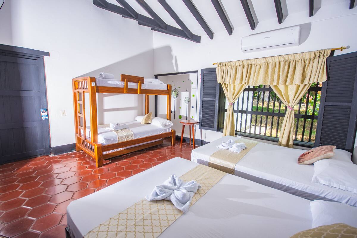 Habitación de hotel con dos camas, una litera de madera, suelo de baldosas, una ventana con cortinas y un equipo de aire acondicionado.