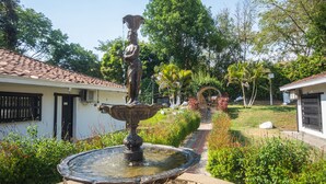 Una fuente con una estatua en un jardín, rodeado de follaje y un sendero que lleva a un edificio con tejado de tejas.