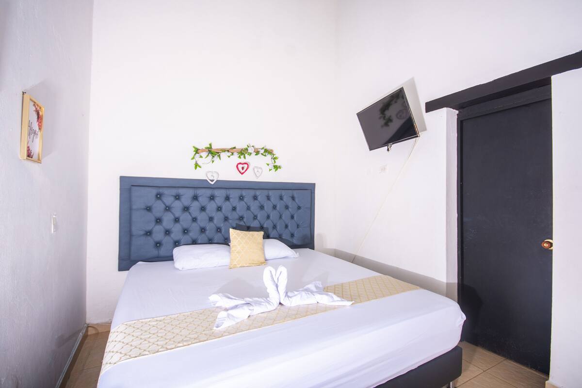 Una cama bien tendida con un cabecero azul, un televisor montado en la pared y una puerta a la derecha.