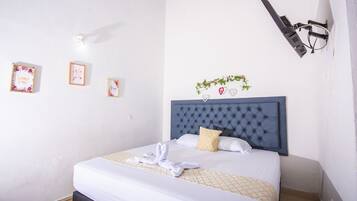 Una cama bien hecha con cabecera capitonada, ropa de cama blanca y una almohada decorativa. Hay cuadros en la pared y una pequeña planta sobre la cama.