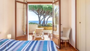Ein Schlafzimmer mit Bett, Stuhl, kleinem Tisch und einem Balkon mit Blick auf das Meer und einen Baum.