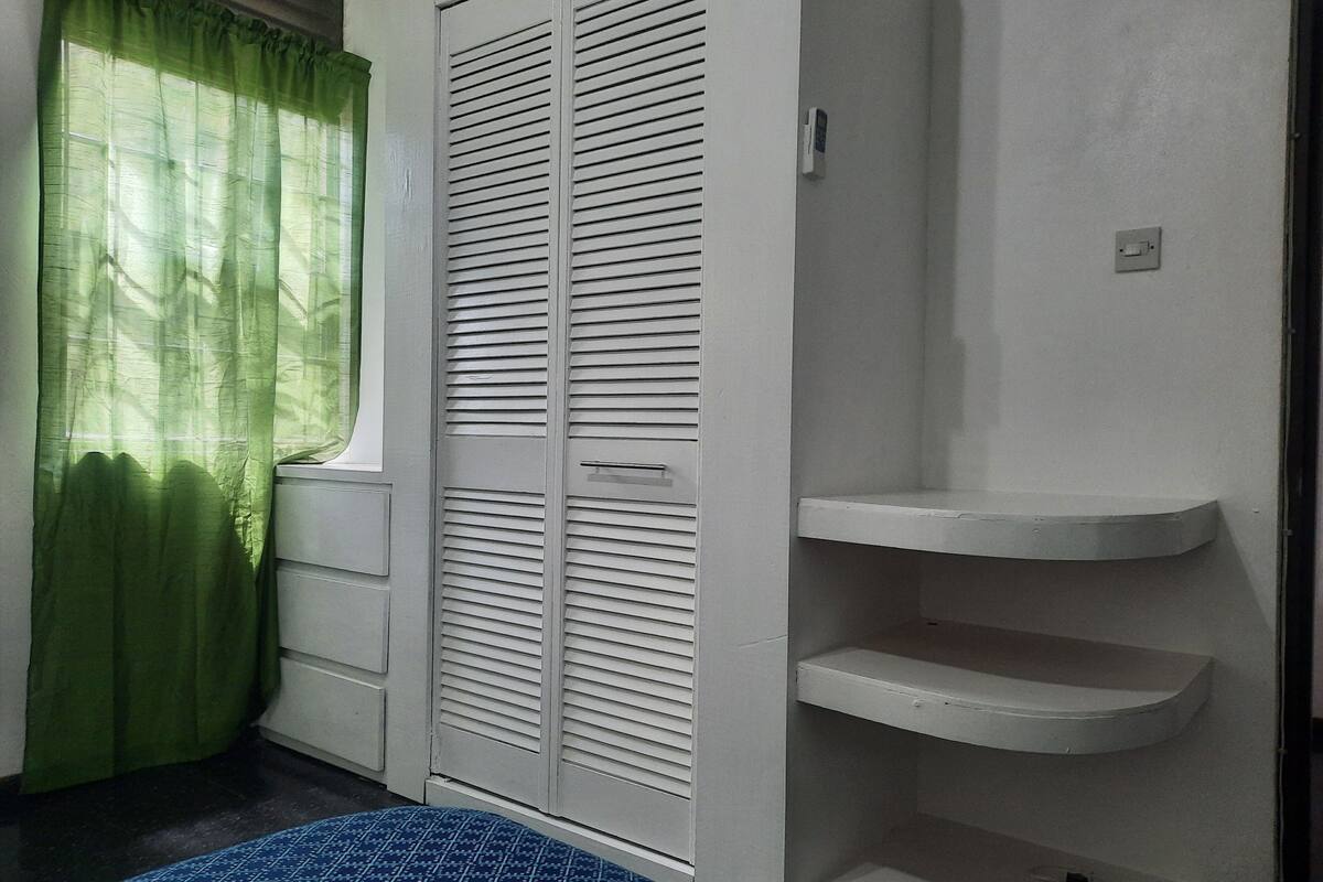 A room with a bed, a wardrobe with horizontal louvered doors, a green curtain, and a white shelf unit.