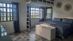 Ein Schlafzimmer mit Etagenbett, einem weißen Nachttisch, einem Bett mit gemustertem Bettbezug und Blick auf Grün hinter den Fenstern.