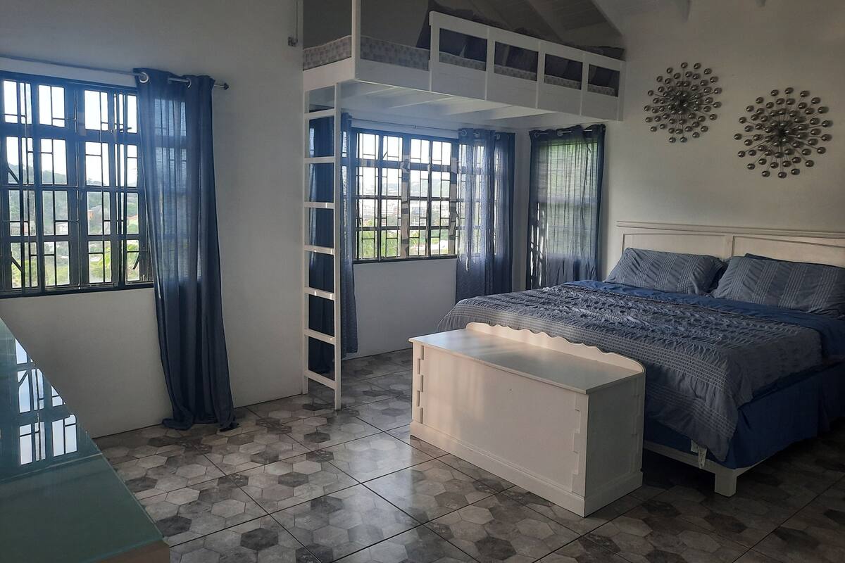 A bedroom with a bunk bed, a white bedside table, a bed with a patterned bedspread, and a view of greenery through the windows.