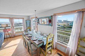 Un coin repas avec une table en bois dressée pour quatre personnes, un lustre et une vue sur la plage grâce à de grandes fenêtres.