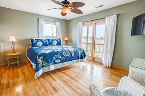 Une chambre avec un lit, deux lampes de chevet, un ventilateur de plafond, une grande fenêtre avec des rideaux et une vue sur la plage.