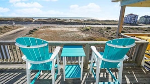 Een houten terras met twee turquoise stoelen en een kleine tafel, met uitzicht op een strand en duinen.