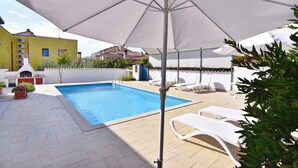 Una piscina con un ombrellone bianco, lettini e una zona barbecue.