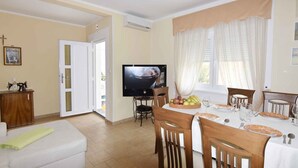 Zona pranzo con TV, sedie in legno, tavolo apparecchiato per due e finestra con tende.