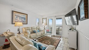 A living room with a beige leather sofa, a small table with a lamp, a framed painting, and a view of the sea through a sliding glass door. - Spacious and newly updated 2nd floor walk-up + Free Attraction Tickets! (North Myrtle Beach)
