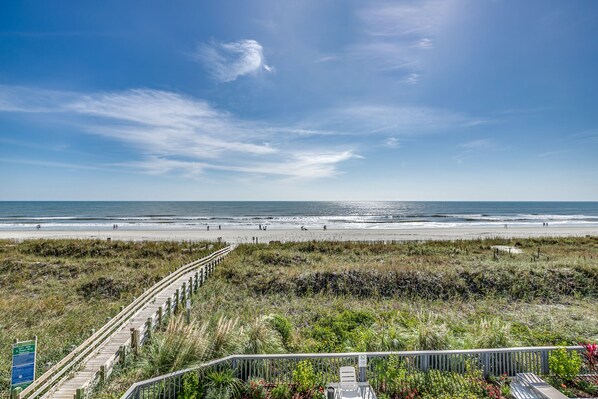 A beach view with a wooden boardwalk leading to the shore, a signboard, and a railing in the foreground.