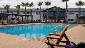Une piscine dotée de chaises longues et d’un espace salon couvert, entourée de palmiers et bordée d’un bâtiment avec des balcons.