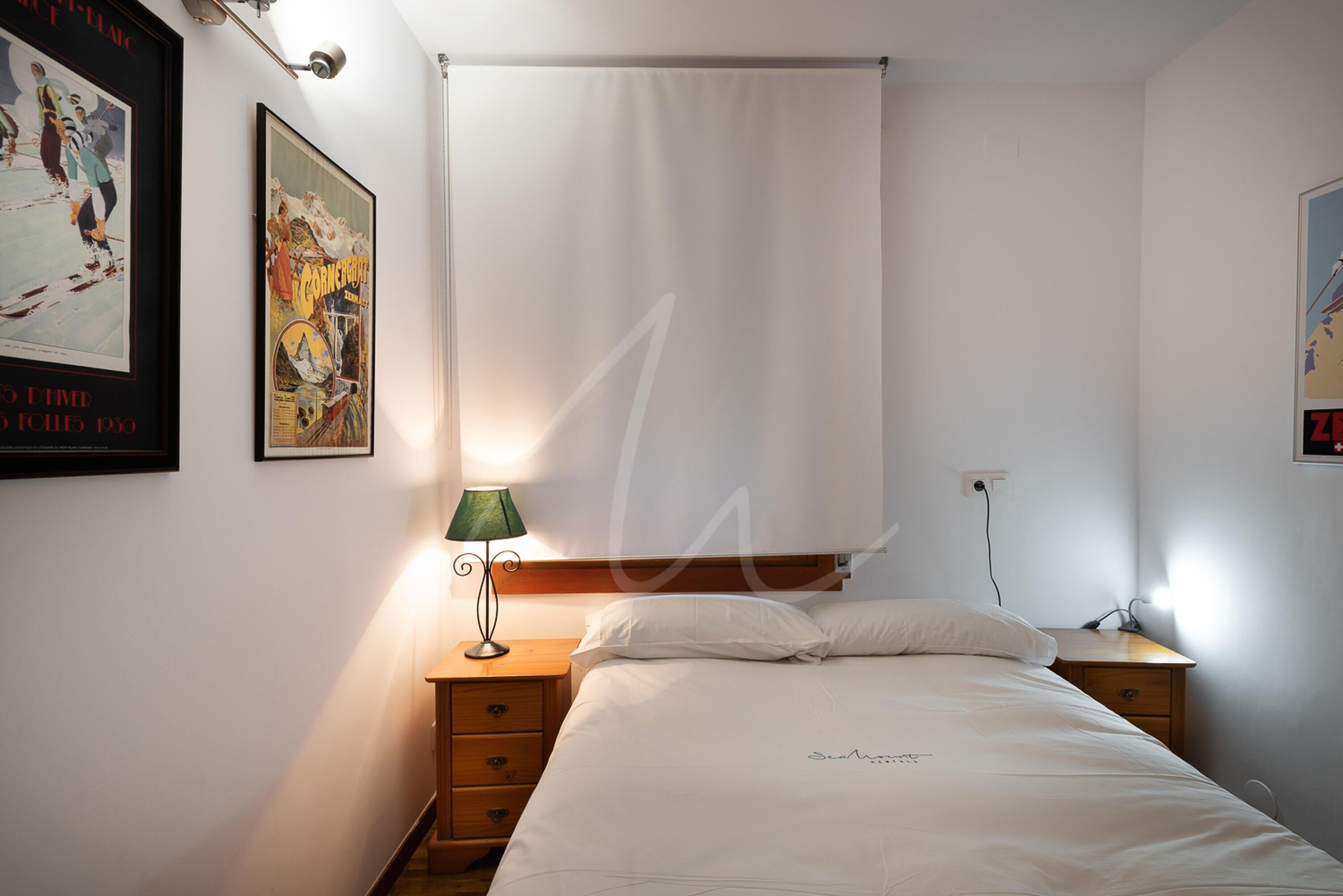 A bedroom with a bed, a nightstand, a lamp, a wall-mounted power outlet, and two posters on the wall.
