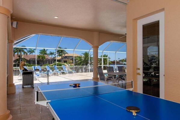 A ping pong table in a covered outdoor area with lounge chairs and a view of palm trees and buildings.