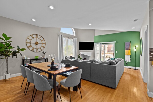 A modern living space with a dining area, a gray sofa, and a large wall clock.