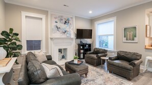 A living room with a fireplace, leather sofas, a flat-screen TV, and a large abstract painting. - Historic 3BR Near Barnes Jewish Hospital | Walkable CWE | Dog-Friendly (St Louis)
