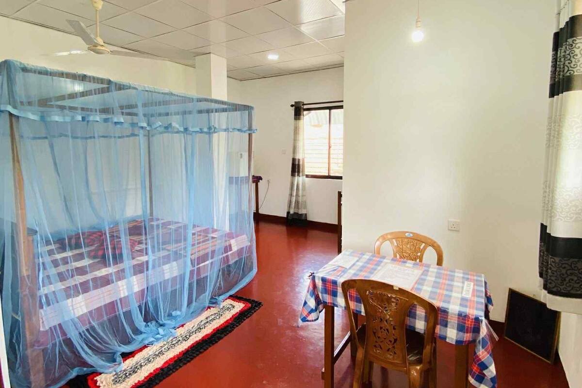 A room with a bed covered by a blue mosquito net, a dining table with a checkered tablecloth, and a chair.