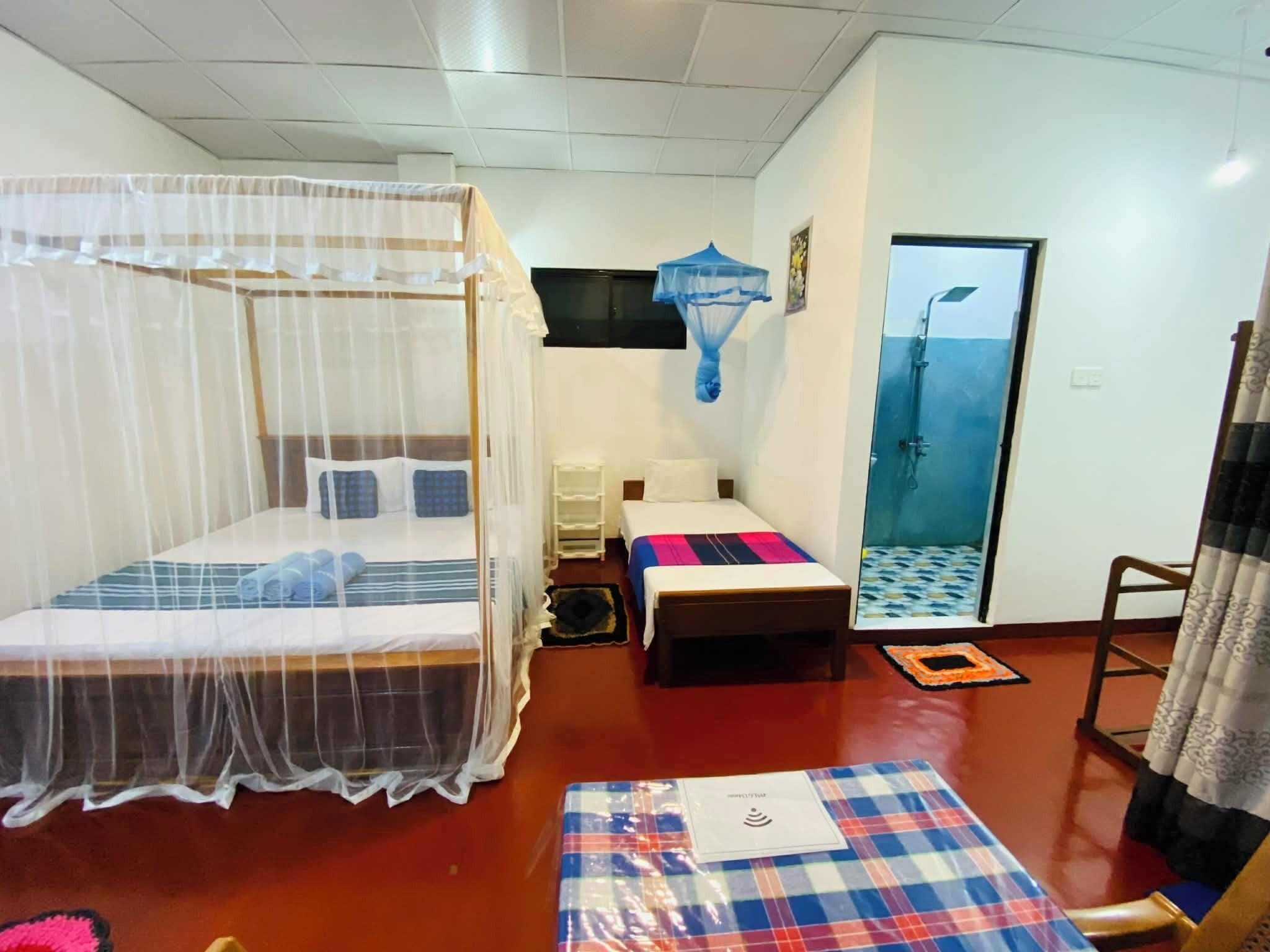 A hotel room with a bed, a small table, a bench, a shower area, and a window.