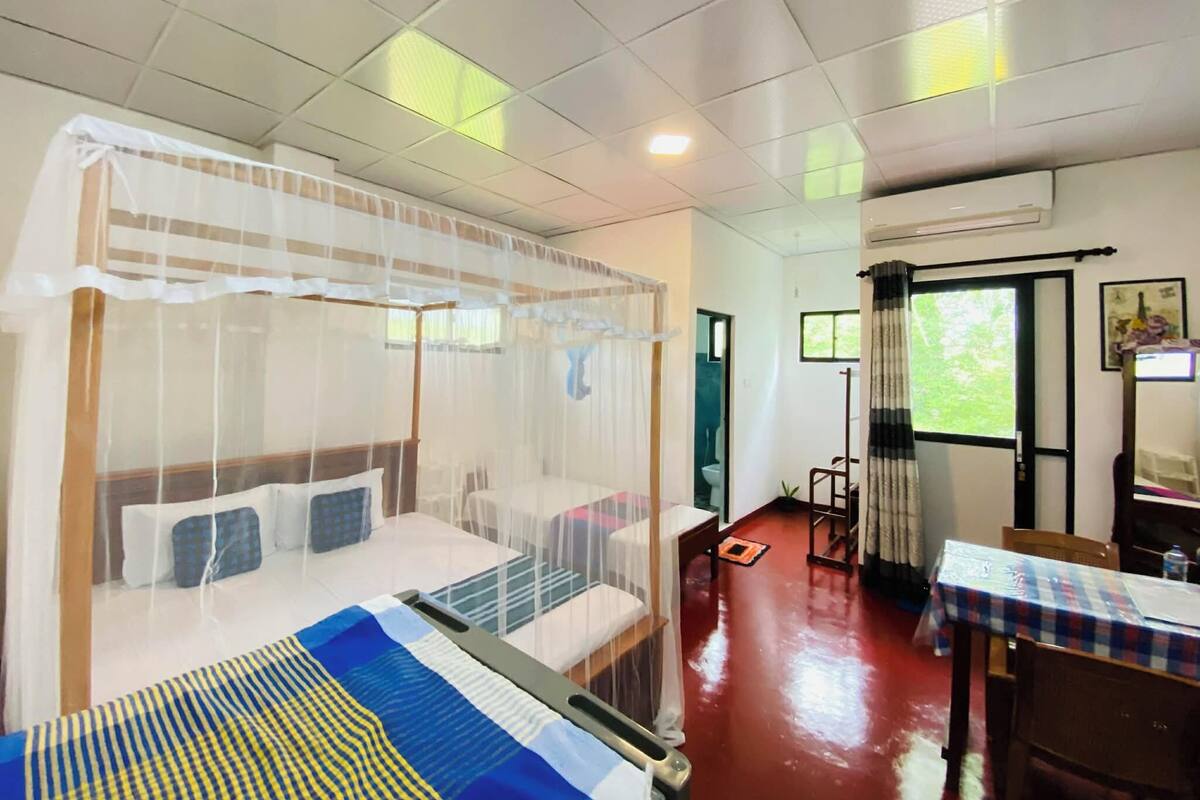 A hotel room with a bed covered by a mosquito net, a small table with a checkered tablecloth, and a window with curtains.