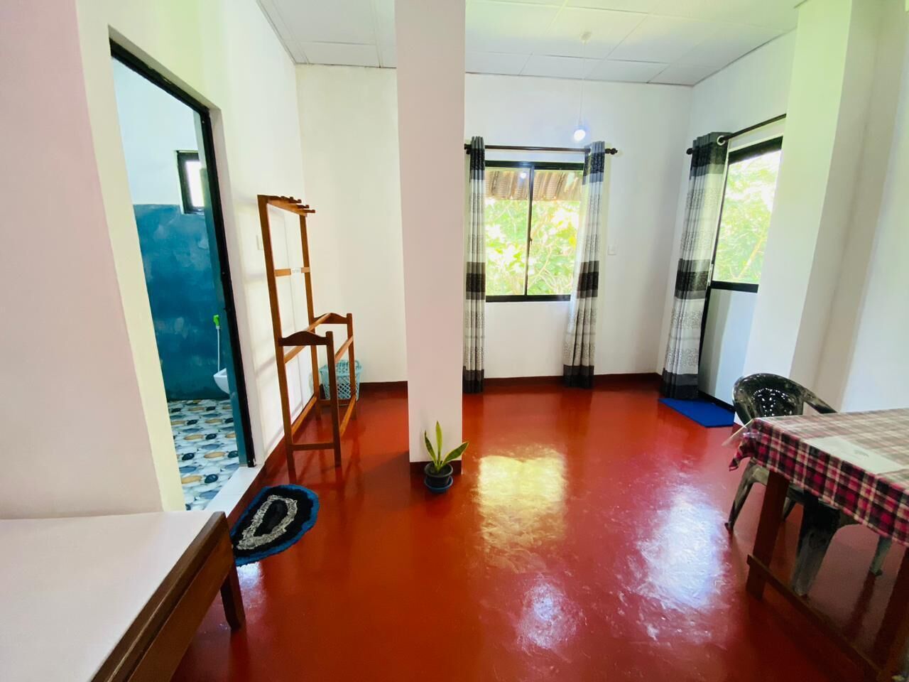 A room with a red floor, a table with a checkered tablecloth, a chair, a wooden ladder, a potted plant, and a window with curtains.