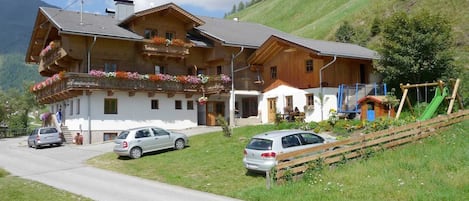A two-story wooden house with a balcony, a car parked in front, and a playground with a slide.