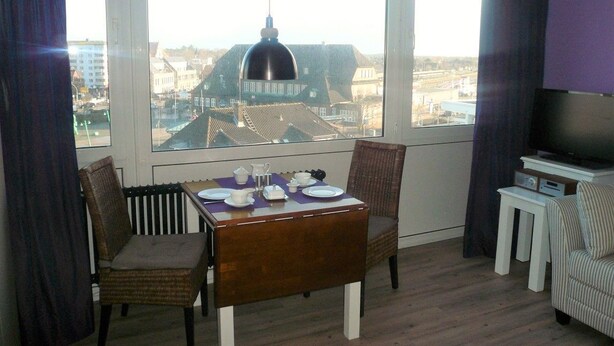 A dining area with a table set for two, a television, and a view of buildings through large windows. - App. Meinecke 29 O, Germany (Sylt)