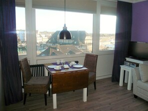 A dining area with a table set for two, a television, and a view of buildings through large windows. - App. Meinecke 29 O, Germany (Sylt)