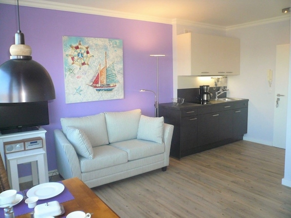 A living room with a sofa, a dining table with plates, a coffee maker, and a painting on the wall. - App. Meinecke 29 O, Germany (Sylt)