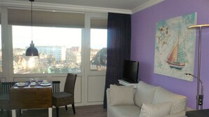 A living room with a dining table, a sofa, a TV, and a large window with a city view. - App. Meinecke 29 O, Germany (Sylt)