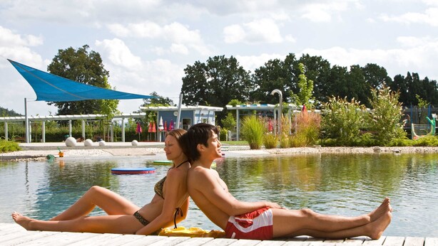 Twee personen ontspannen op een houten steiger aan een kalm waterlichaam, met een blauw zonnescherm erboven en een gebouw op de achtergrond.
