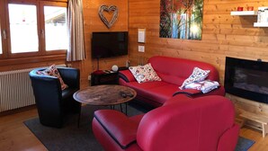 Una sala de estar acogedora con un sofá rojo, un sillón negro y una mesita de centro. Hay un televisor de pantalla plana montado en la pared, una decoración con forma de corazón y un cuadro con árboles.