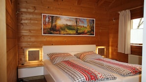Un dormitorio de cabaña de madera con una cama, ropa de cama cuadriculada, un cuadro de un paisaje y una mesita de noche pequeña.