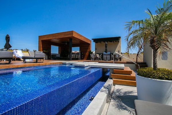 A swimming pool with a wooden deck, lounge chairs, and a covered seating area.