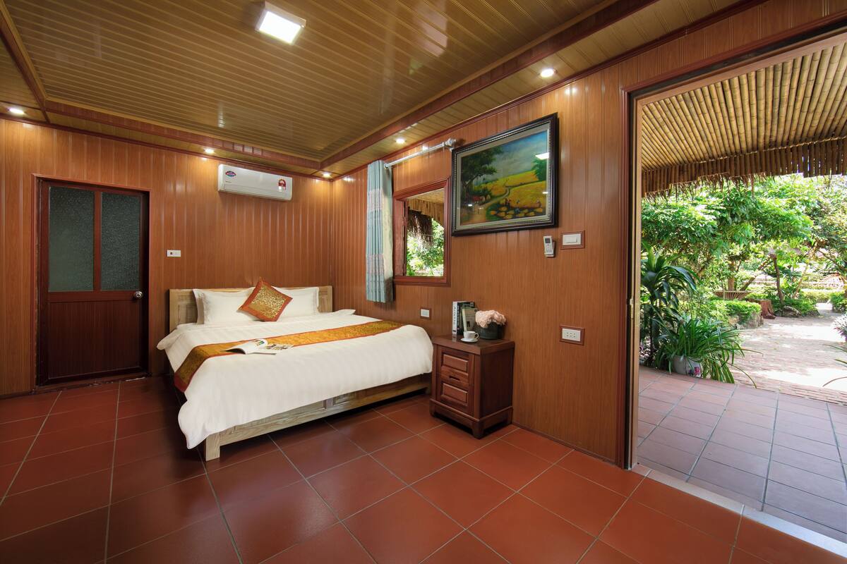 A hotel room with a bed, wooden walls, a painting, and a view of greenery.