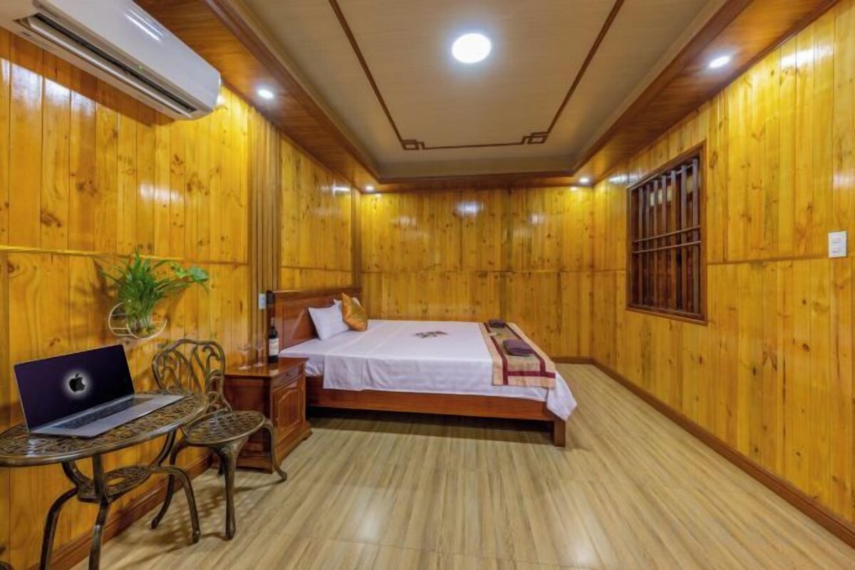 Habitación con paneles de madera, una cama, un escritorio con una computadora portátil y una planta en maceta.