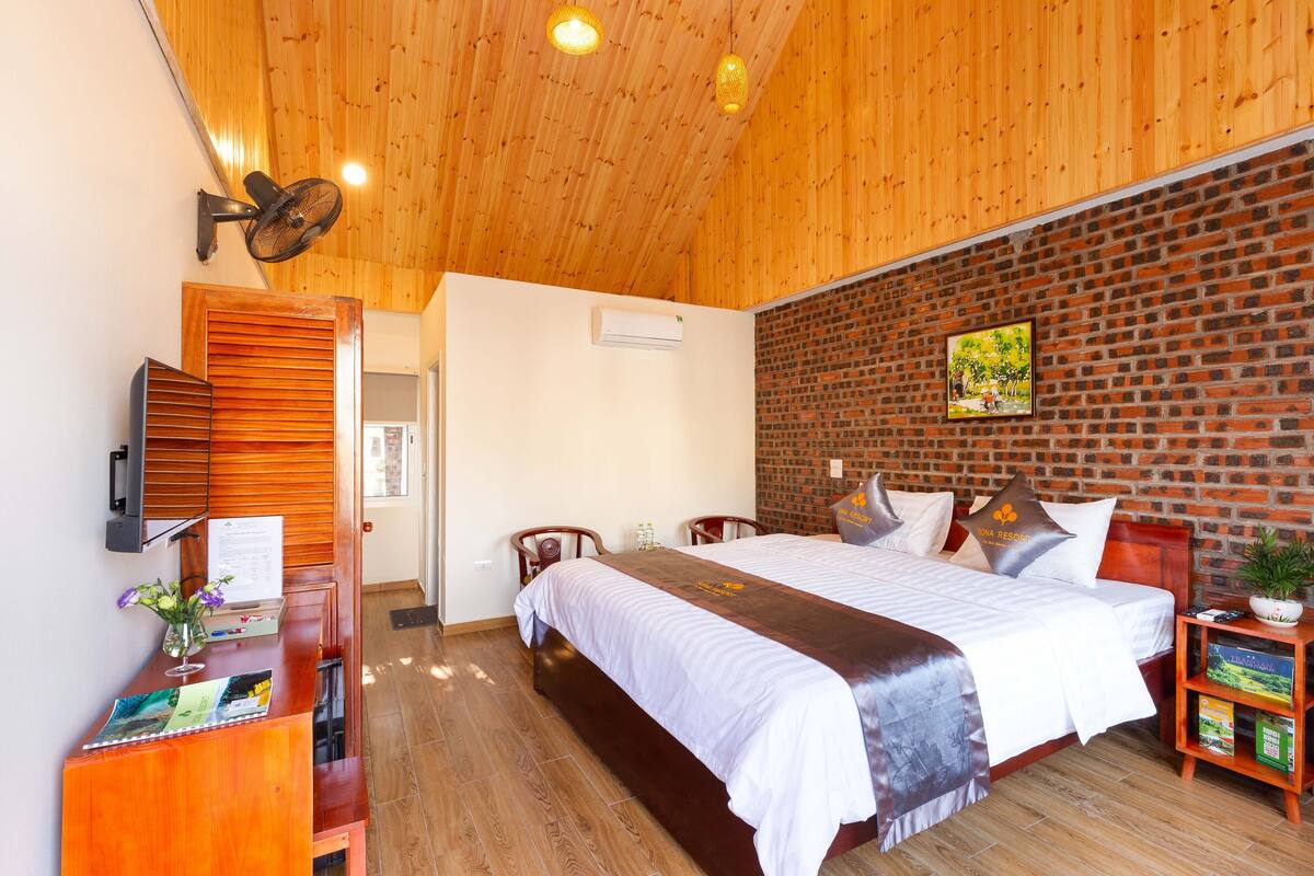 Ein Hotelzimmer mit Holzboden, einer Ziegelwand, einem großen Bett mit Kissen, einem Schreibtisch mit Computer und einer kleinen Pflanze.