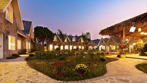 A stone-paved courtyard with a thatched-roof structure, surrounded by well-maintained gardens and a row of traditional-style buildings with visible windows and doors.