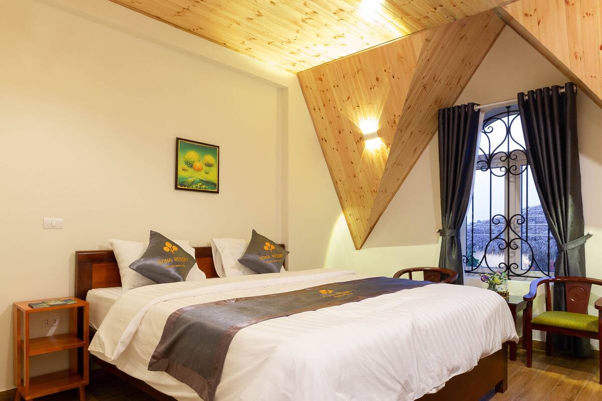 Ein Schlafzimmer mit Holzbalkendecke, einem Bett mit weißer Bettwäsche, einem Nachttisch, einem Sessel und einem Fenster mit Vorhängen.