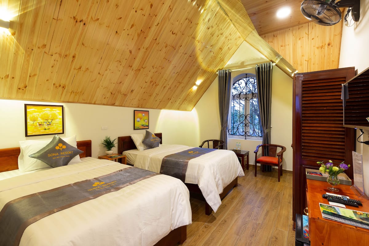 Ein Hotelzimmer mit zwei Betten, Holzboden, einem Fenster mit Vorhängen, einem Schreibtisch mit einer Vase und einem Stuhl.