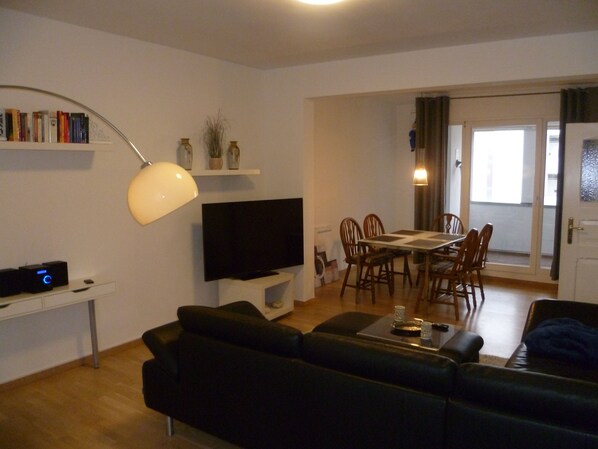 A living room with a black leather sofa, a flat-screen TV, a dining table with chairs, and a bookshelf.